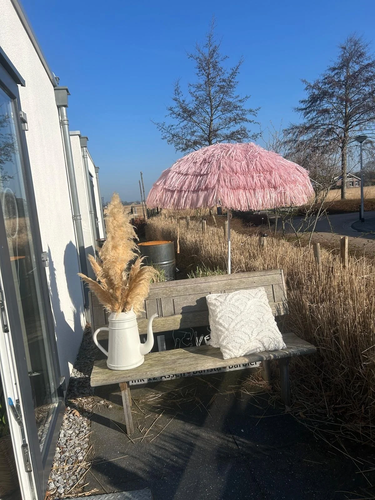 Houten bankje met pampasgras in een gieter en een roze parasol